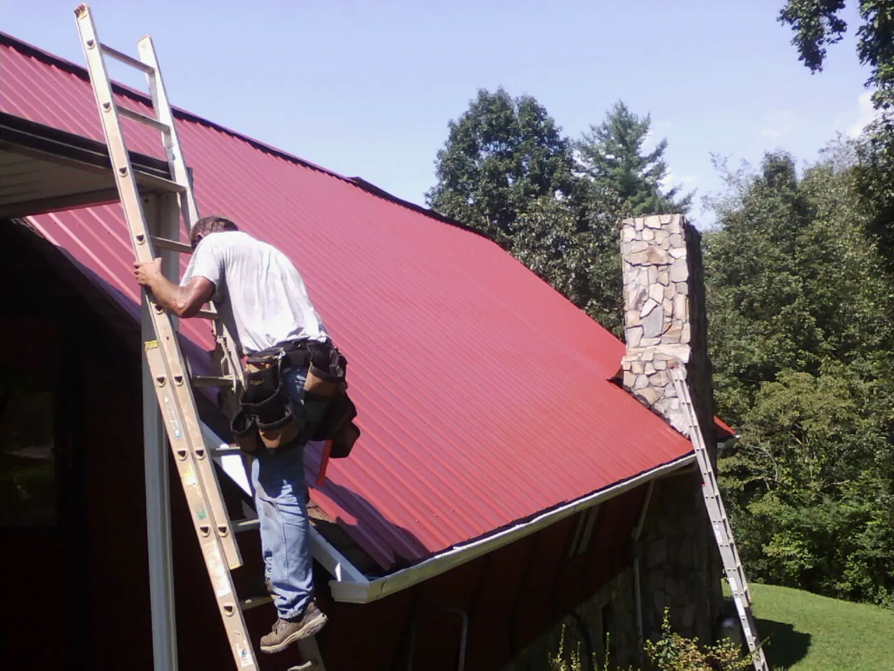 Roofer on a ladder beside a red metal roof and stone chimney — Soffit & Fascia Repair by Roofing Brothers in Burket